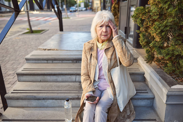 Sad woman with bottle of water sitting on the steps near the building - 写真・画像