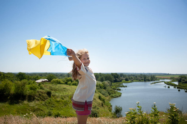 Ευτυχισμένο χαμογελαστό κορίτσι από την Ουκρανία στη Vyshyvanka με κίτρινη και μπλε σημαία σε ένα βουνό στο βάθος των αγρών και ενός ποταμού. Ημέρα Ανεξαρτησίας της Ουκρανίας. Εθνική Σημαία της Ουκρανίας. - Φωτογραφία, εικόνα