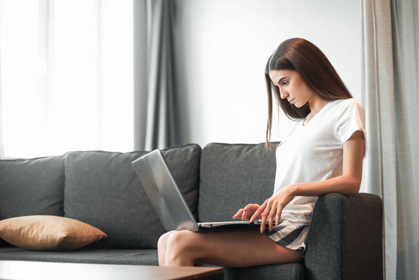 Young woman with modern laptop sitting on sofa at home. Happy girl browsing through the internet during free time..  - Фото, изображение