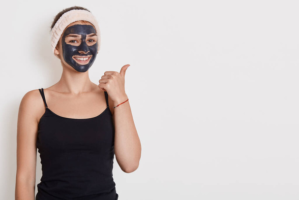 Photo of smiling woman wears black t shirt and hair band with facial mask, has beauty procedures at home, positive expression, points aside with thumb isolated over white wall. - Foto, Imagem