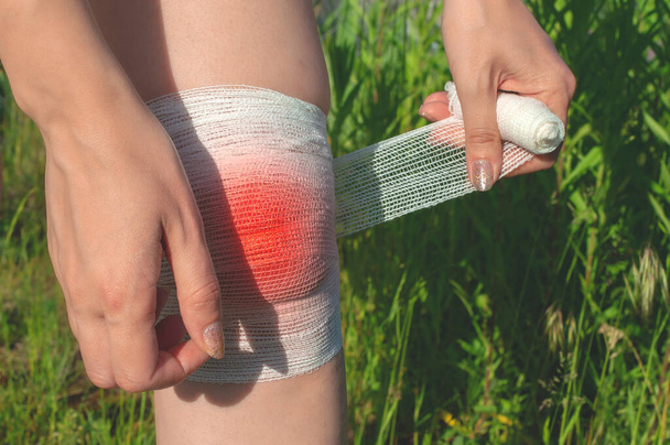 Een jonge vrouw buiten in het midden van een veld doet zelfstandig een verband om haar gewonde knie. Close-up van het epicentrum van een blauwe plek - Foto, afbeelding