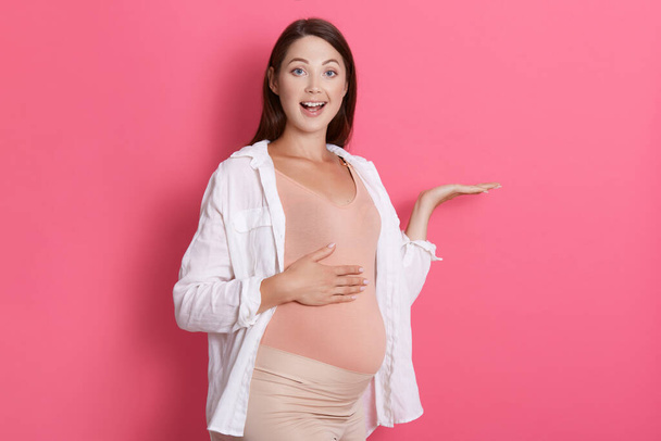 Young beautiful girl expecting baby posing isolated over pink background pointing aside with palms, showing copy space, presenting advertisement, being astonished. - Fotografie, Obrázek