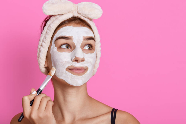 Girl receiving cosmetic white facial mask, looking dreamily aside, wearing hairband with bow, charming lady doing anti aging procedures at home in morning. - Φωτογραφία, εικόνα