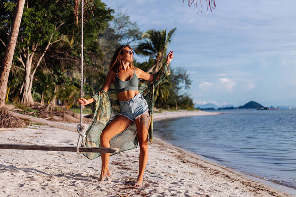 Fit tanned good looking tattooed caucasian woman in denim shorts and green trendy crop top, tropical print cape, on exotic beach on swing at sunset - Photo, Image