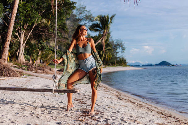 Fit tanned good looking tattooed caucasian woman in denim shorts and green trendy crop top, tropical print cape, on exotic beach on swing at sunset - Φωτογραφία, εικόνα