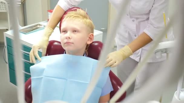Dişçi konsepti. Çocuk dişçi muayenehanesinde dişçi koltuğunda oturan çocuk hasta. Peçete yerleştirme ve onarım asistanı. Diş kliniğinde tedavi gören bir çocuk. 4k video - Video, Çekim