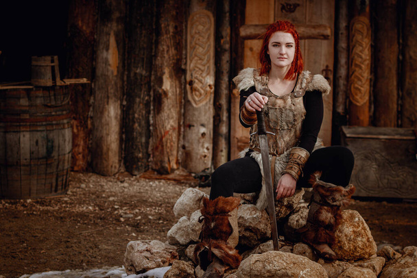 赤い髪のバイキングの女の子。剣の手に敵と戦うために。中世の風景の再構築。大規模なバイキング村を背景に. - 写真・画像