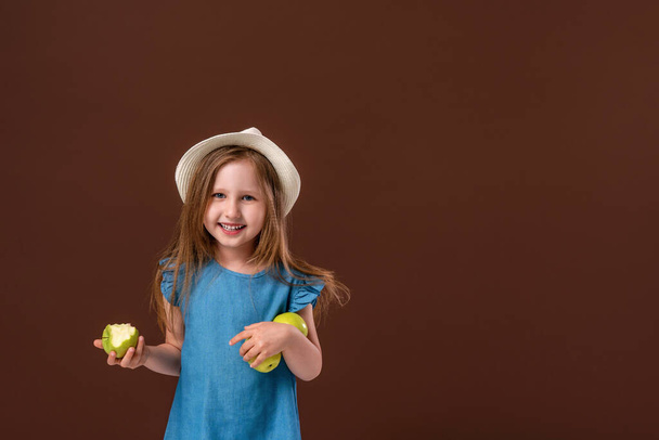Portrait of a cute little baby girl holding an Apple on a brown background. The child is wearing a straw hat and eating an Apple. Gathered the autumn harvest and eats fruit. The autumn harvest. - Photo, Image