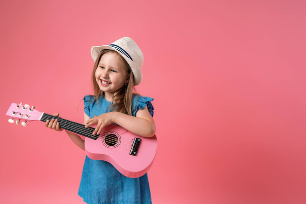 cheerful little Caucasian girl of 4 years old in a short denim dress and hat plays a pink ukulele guitar. A child plays a musical instrument and sings on a pink background. - Valokuva, kuva