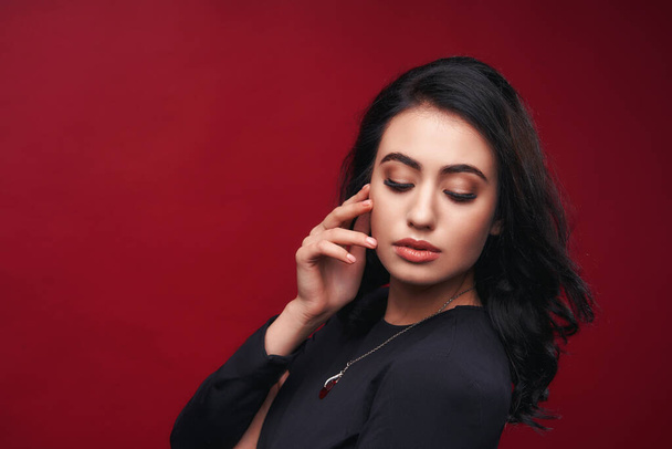 Portrait attractive, sensual woman with evening makeup and parted lips, black hair loose, looking over her shoulder, posing indoors against red background Studio. classical concept of makeup. - Foto, Bild