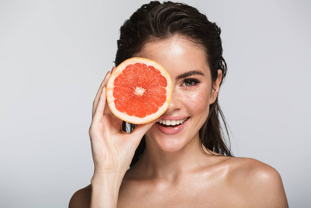Beauty portrait of an attractive smiling sensual young woman with wet brunette long hair standing isolated over gray background, showing halved grapefruit, posing - Foto, immagini