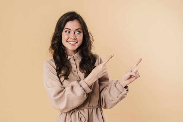 Image of cheerful attractive woman smiling and pointing fingers aside isolated over beige background - Foto, imagen
