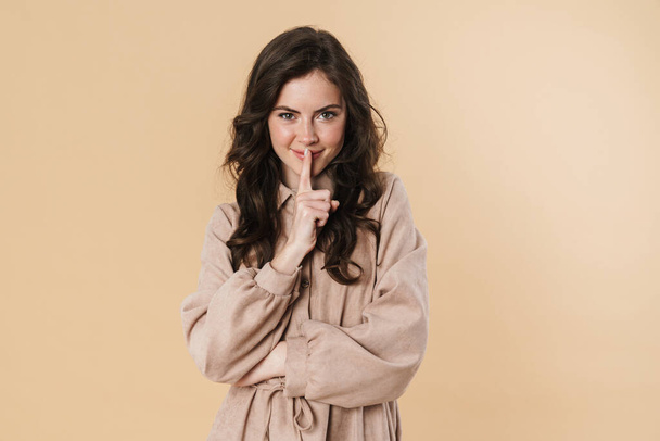 Image of pleased nice woman showing silence gesture and looking at camera isolated over beige background - Фото, изображение