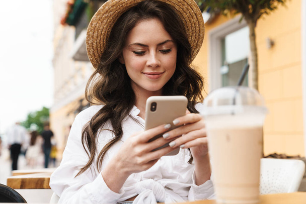 Portrait of young smiling woman using cellphone while drinking milkshake in cozy cafe outdoors - Photo, Image