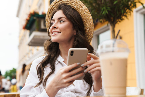 Muotokuva nuori hymyilevä nainen käyttää matkapuhelinta juodessaan pirtelöä viihtyisässä kahvilassa ulkona - Valokuva, kuva