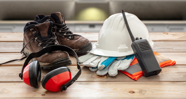 Work safety protection equipment. Industrial protective gear on wooden table, blur construction site background. Hardhat, boots, earmuffs, walkie talkie, gloves and vest - Foto, afbeelding