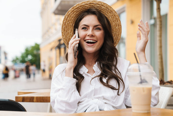 Portrait of young laughing woman talking on cellphone while drinking milkshake in cozy cafe outdoors - Foto, Bild