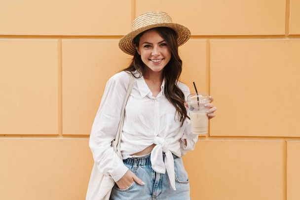 Portrait of young happy woman in straw hat drinking milkshake and smiling while walking on city street - Foto, Bild