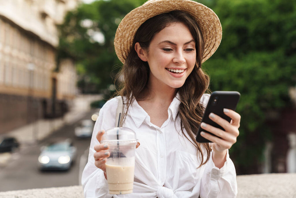 Portrait of young joyful woman using cellphone and drinking milkshake while walking on city street - Photo, Image