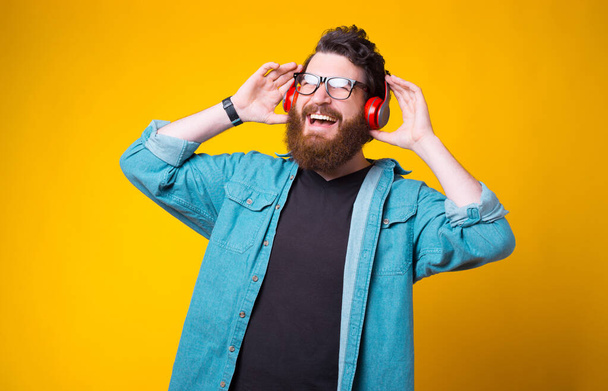 Cheerful bearded hipster is listening to the music through headphones over yellow background - Фото, зображення