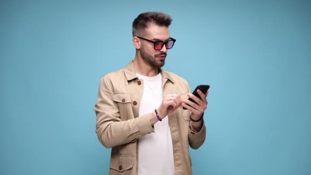 happy casual man in jacket holding phone and reading emails, covering mouth with hand and dancing, pointing finger to phone and making call me sign, moving and gesturing on blue background - Footage, Video