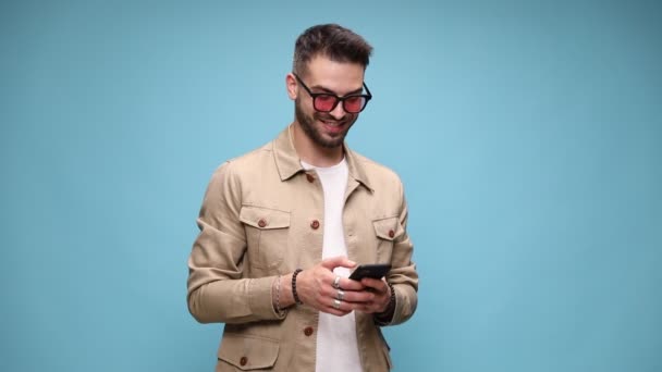 happy young guy in jacket wearing sunglasses, looking up and down, writing messages and smiling, pointing finger and laughing on blue background - Footage, Video