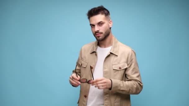 sexy young man in jacket straightening the temples and putting glasses on, adjusting jacket and holding hand behind neck in a fashion pose on blue background - Footage, Video