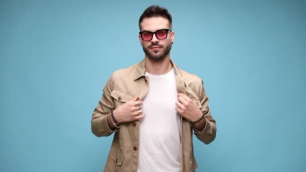 sebevědomý mladý ležérní muž nosí brýle, upravuje bundu a drží ruku v kapse, stojí v módní póze na modrém pozadí - Záběry, video