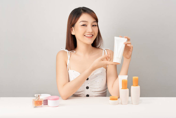 Jovem mulher asiática segurando creme de cuidados da pele com rosto sorridente isolado no fundo branco
. - Foto, Imagem