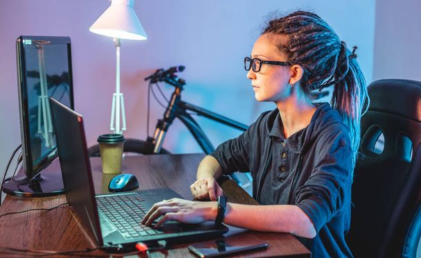 Σύγχρονη νεαρή γυναίκα προγραμματιστής γράφει κώδικα προγράμματος σε ένα φορητό υπολογιστή στο σπίτι. Απομακρυσμένη εργασία στο επάγγελμα της πληροφορικής. Η έννοια της ανάπτυξης λογισμικού. - Φωτογραφία, εικόνα
