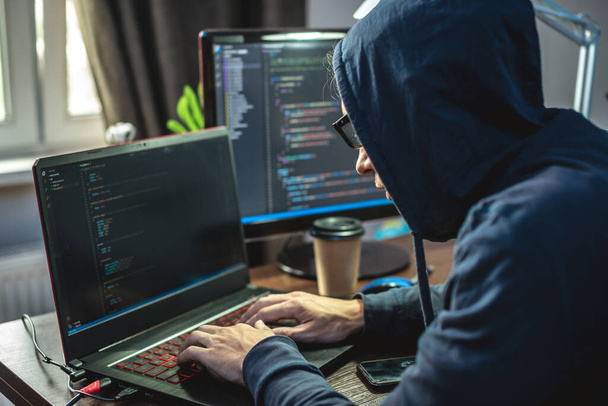 Man hacker in de motorkap programmeert virus code op het toetsenbord voor de laptop. Het concept van internetfraude en het hacken van persoonsgegevens - Foto, afbeelding