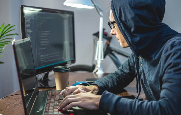 フードの男のハッカーはラップトップでウイルスコードをプログラミングしてる。インターネット詐欺や個人データハッキングの概念 - 写真・画像