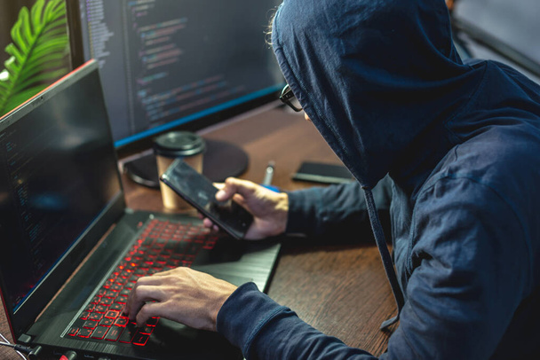 フードの男のハッカーは、ラップトップのキーボードでウイルスコードをプログラミングしています。インターネット詐欺や個人データハッキングの概念 - 写真・画像