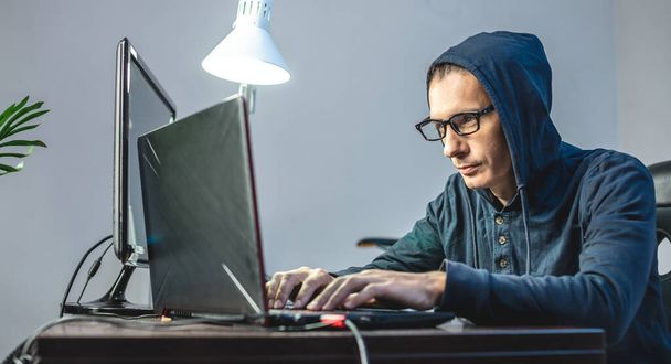 Man hacker in the hood is programming virus code at the keyboard for the laptop. The concept of Internet fraud and personal data hacking - 写真・画像