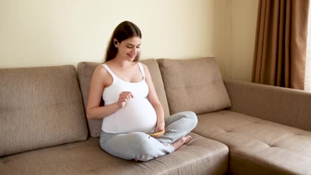 pregnant woman applying headphones in the sofa. Expectant mother listening music and dancing in light bedroom. Pregnant female giving unborn listening music at home - Filmagem, Vídeo