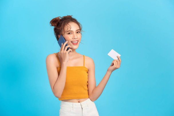 Portret van tevreden vrouw praten op mobiele telefoon en houden credit card in de hand geïsoleerd over blauwe achtergrond - Foto, afbeelding