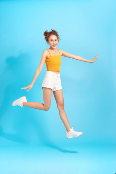 Full length image of happy active asian woman jumping on blue background. Summer trendy shorts and crop top. - 写真・画像