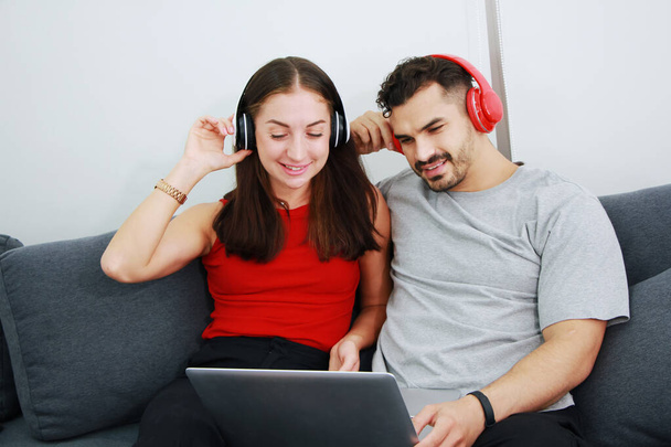 Lächelndes junges kaukasisches Liebespaar, das mit Kopfhörern und Laptop gemeinsam im Wohnzimmer Online-Musik hört. Lebensstil und Beziehung des Ehelebens im Urlaub. - Foto, Bild