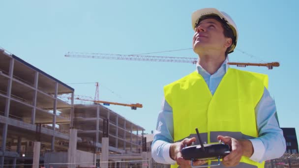 Drone operator holding remote controller. Professional builder in helmet and vest standing in front of construction site. Office building and crane background. Business, real estate and investment - 映像、動画