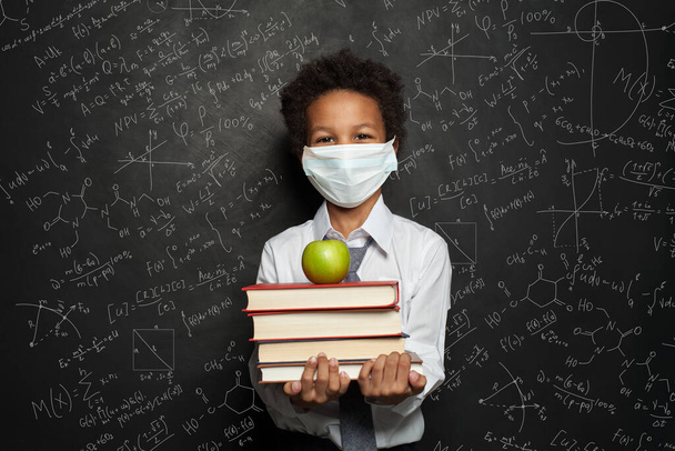 Happy child in medical protective face mask holding books and green apple on blackboard background - Fotoğraf, Görsel