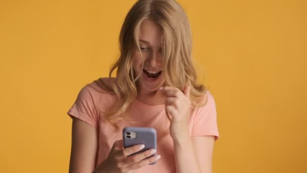 Young excited blond girl intently using smartphone on camera over colorful background. Wow expression - Πλάνα, βίντεο