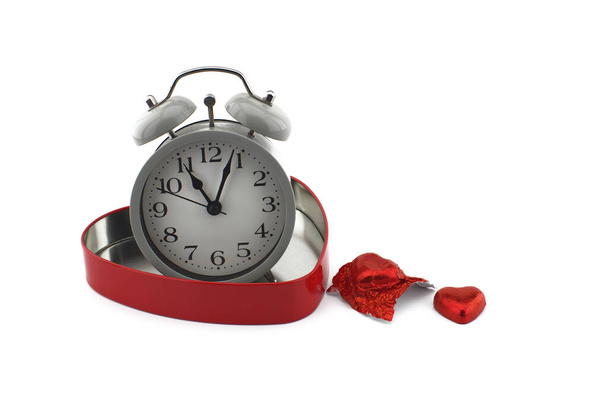 Kırmızı kalp şeklinde bir kutuda çalar saat. Yanında çikolata şekeri ve beyaz bir arka plan. Sevgililer Günü için aşk, birliktelik ve romantizmin simgesi. - Fotoğraf, Görsel