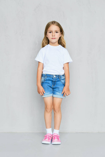 Little child girl in white t-shirt, jeans shorts and pink sneakers standing straight near the wall - Foto, immagini