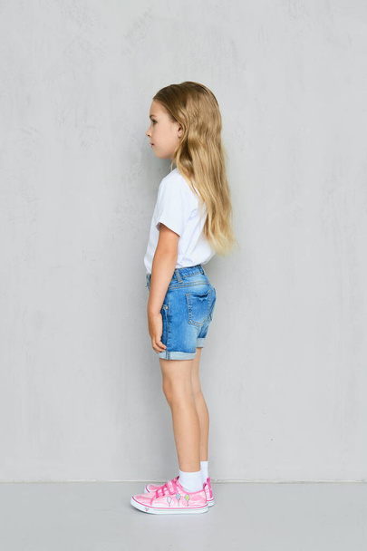 Little child girl in white t-shirt, jeans shorts and pink sneakers standing in profile near the wall - Fotoğraf, Görsel