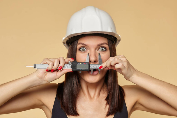 woman in a White protective construction helmet holds a caliper. performs the measurement of the nose. a fun concept for building advertising - Foto, afbeelding