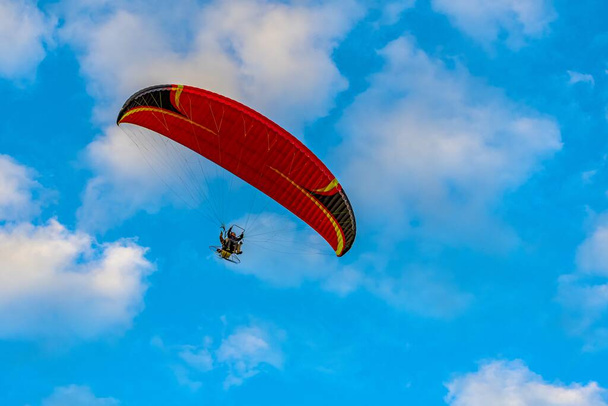 Red dome of a motor paraglider flying on a background of blue cloudy summer sky. Tandem on a paraglider high in the sky. - Foto, afbeelding