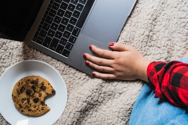 A horizontal shot of a female sitting on a blanket with her laptop and a cookie - Φωτογραφία, εικόνα