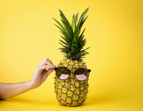 A person's hand putting a sunglasses on a pineapple against a yellow background - travel, summer, vacation concept - Fotó, kép