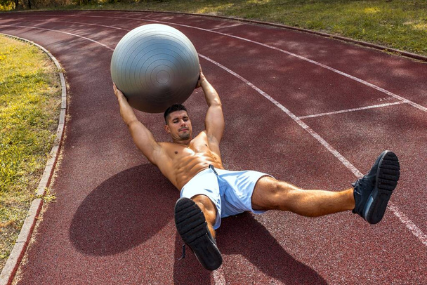 A fit shirtless man exercising with a gray ball on the floor in a park - Foto, Imagen