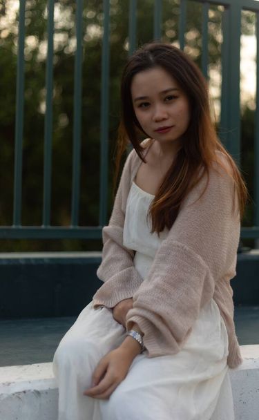 An attractive Asian young female with long hair wearing a white dress with a jacket sitting near a metal fence - Foto, afbeelding